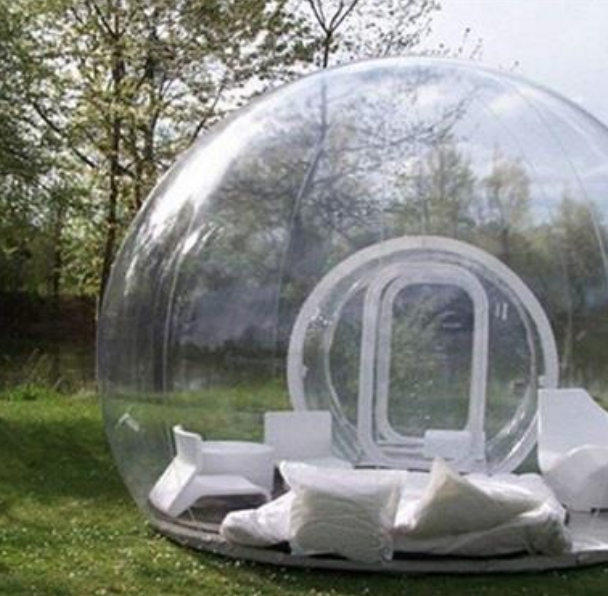 Transparente Kuppel-Lounge auf Wiese mit weißen Sitzmöbeln, Kissen und Bäumen im Hintergrund.